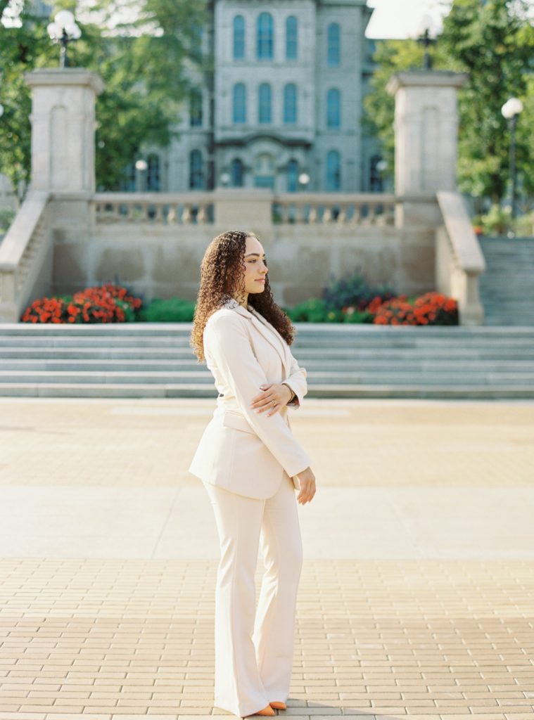 Newhouse Syracuse University student poses in front of main campus during grad session, wearing all ivory suit