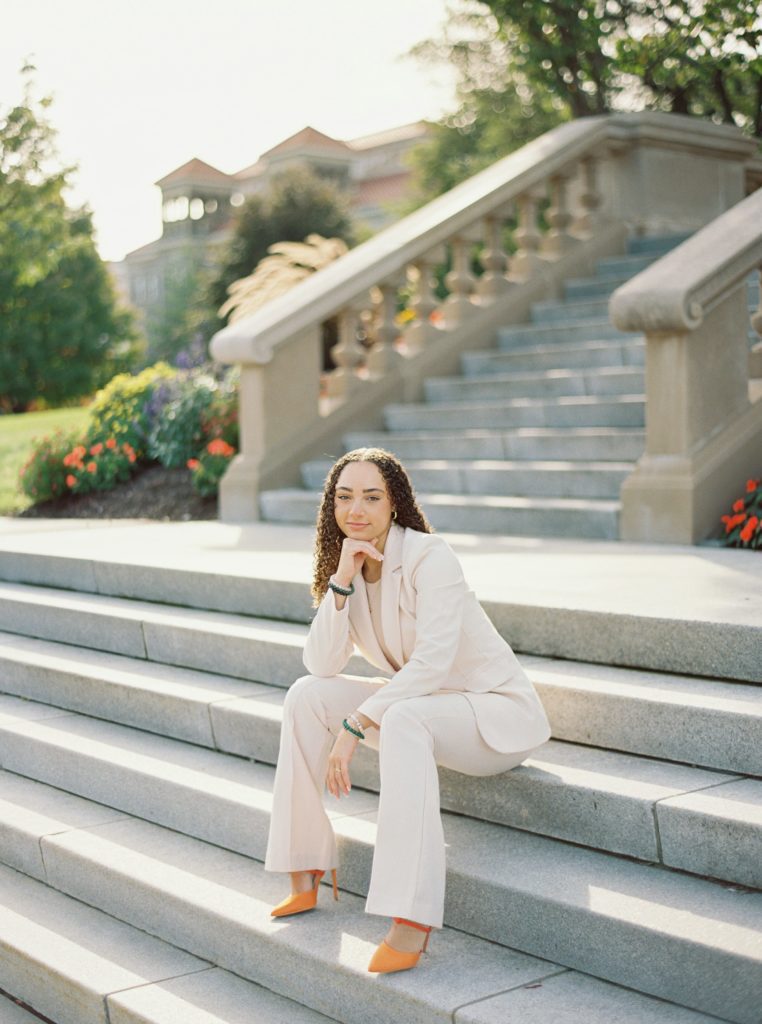 Woman sits on SU steps and looks right into the camera, wearing orange heels and an ivory suit